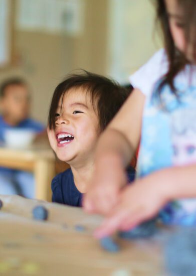 Children in a childcare centre classroom. Child in the foreground doing sensory art play with clay, beside a smiling child. Another child in the background.
