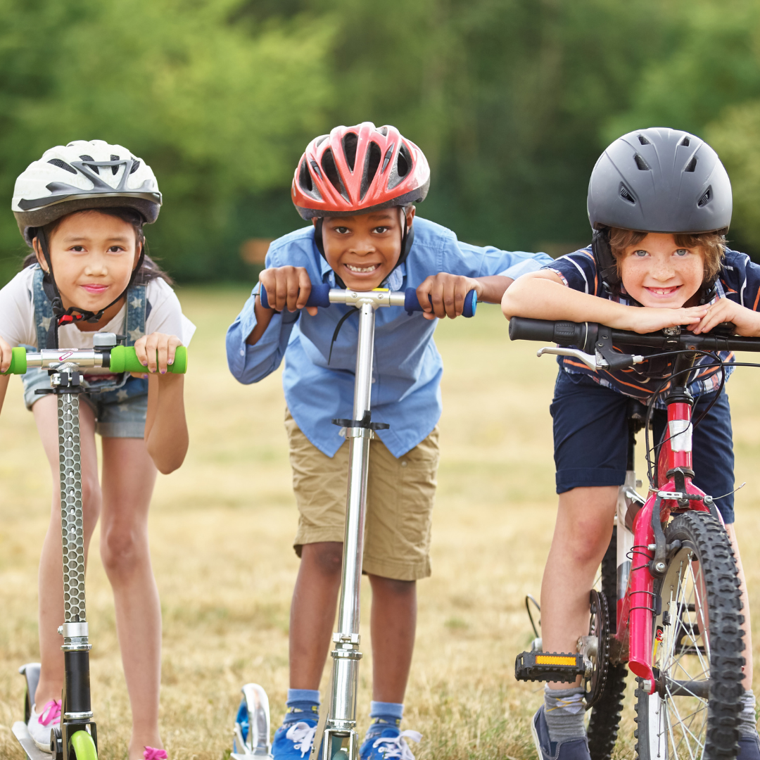 Three kids wearing helmets: 2 on scooters, 1 on bike