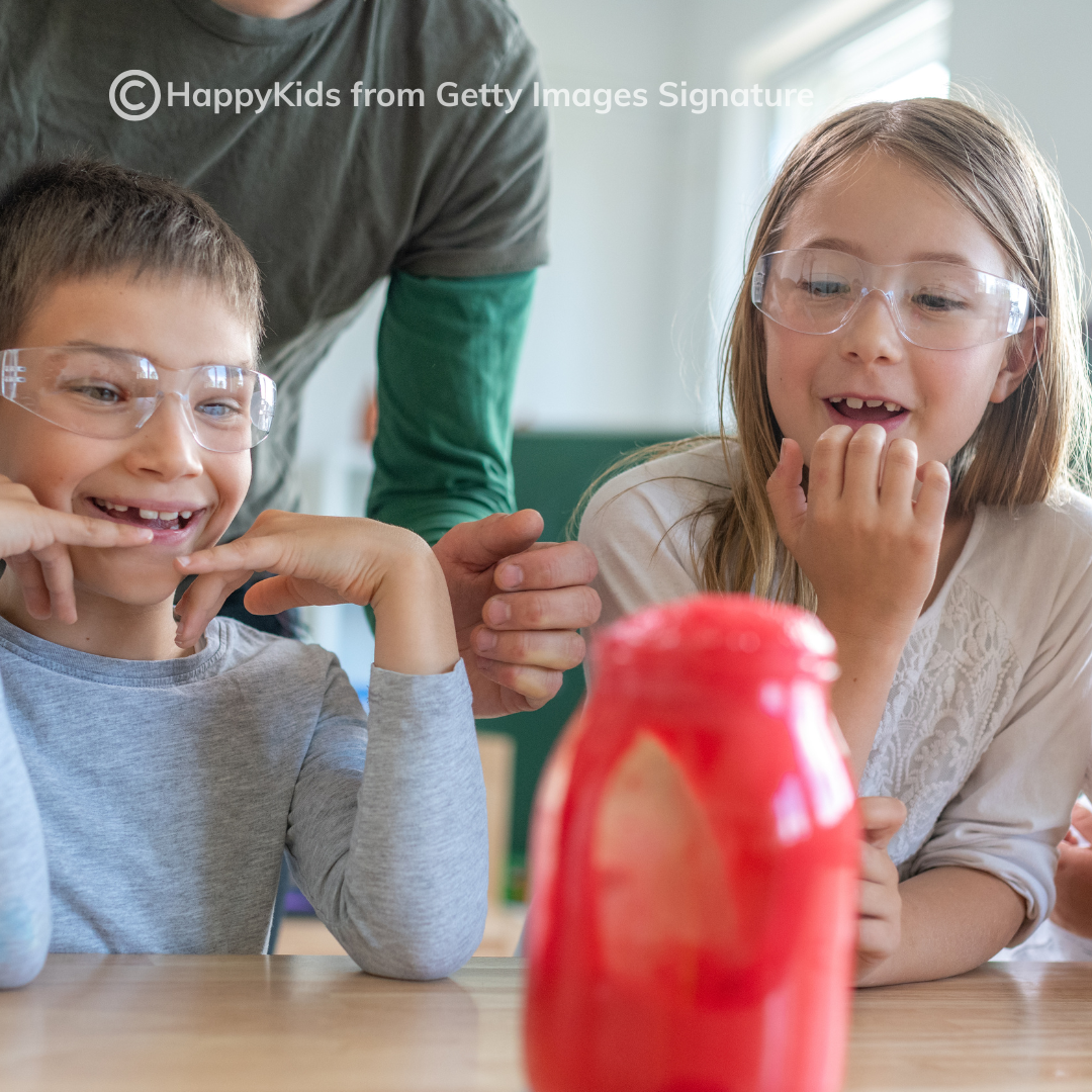 School age children wearing goggles and doing science experiment with adult in the background