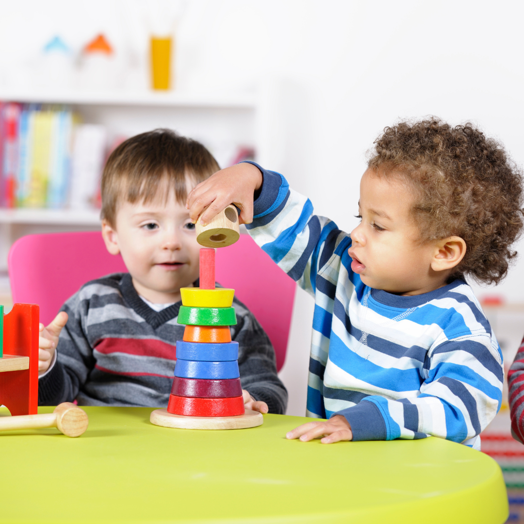 Two toddlers sitting on chairs and playing with a stacking toy at a table