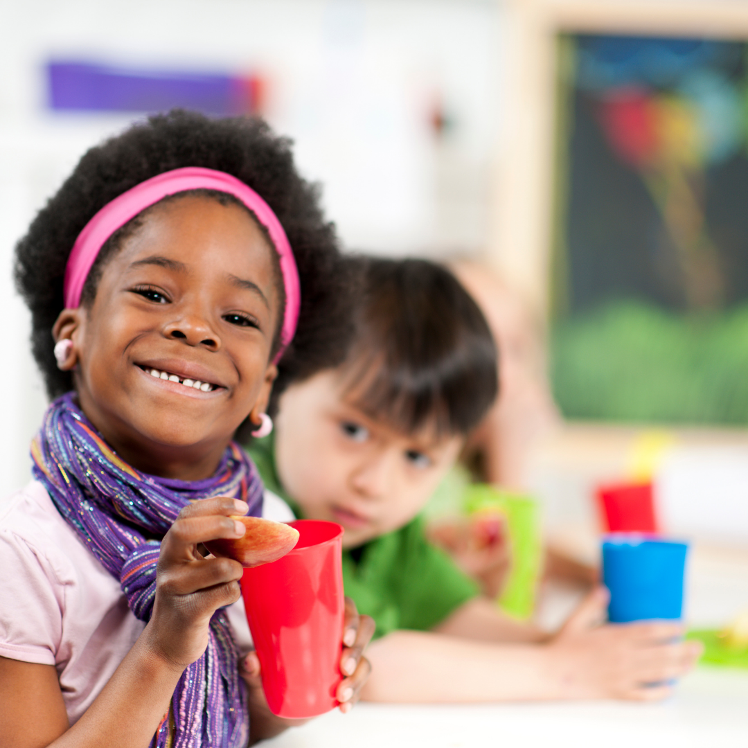 Happy child holding plastic cup, at a classroom table, beside other child