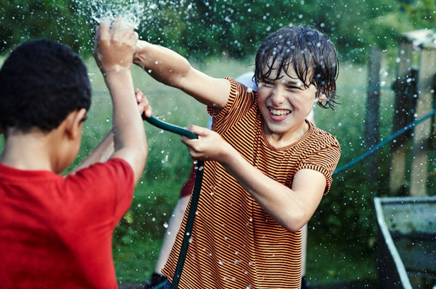 Two children playing with water hose. Children are facing each other and only one child's face is visible. Lots of water is spraying out of the hose.