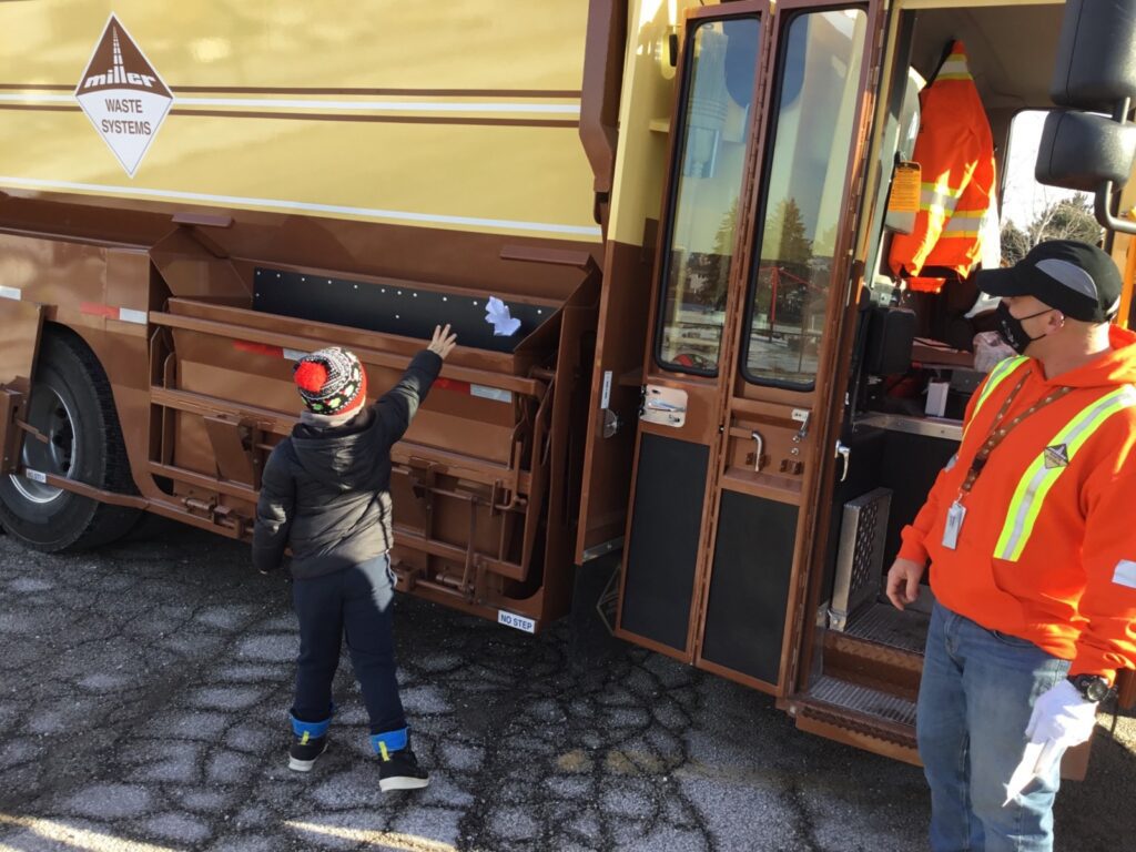 A child throws a piece of paper into the side compartment of a garbage truck as worker stands by