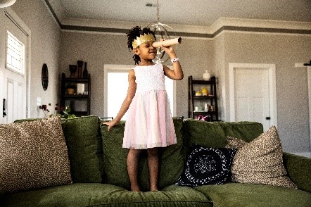 Child standing on a sofa, wearing a crown, a dress, and looking through a paper roll like it is a telescope