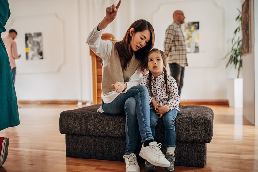 An adult and child sitting together in an art gallery. Adult is pointing to something and child is looking at it. Other adults standing in the background looking at art.