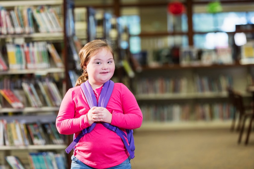 A smiling child standing with a backpack in a library