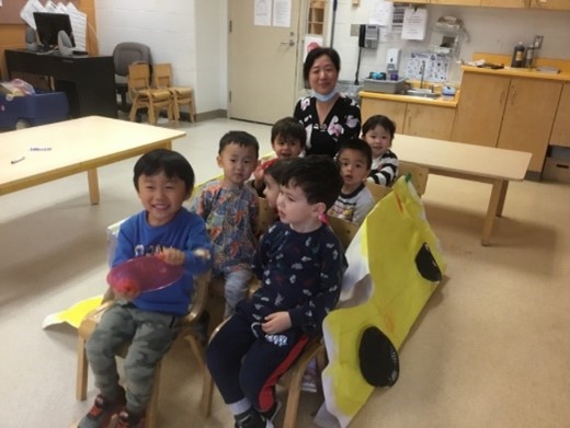An educator and six children in a childcare classroom, pretending to be on a bus (made out of chairs and a painted picture of a bus)