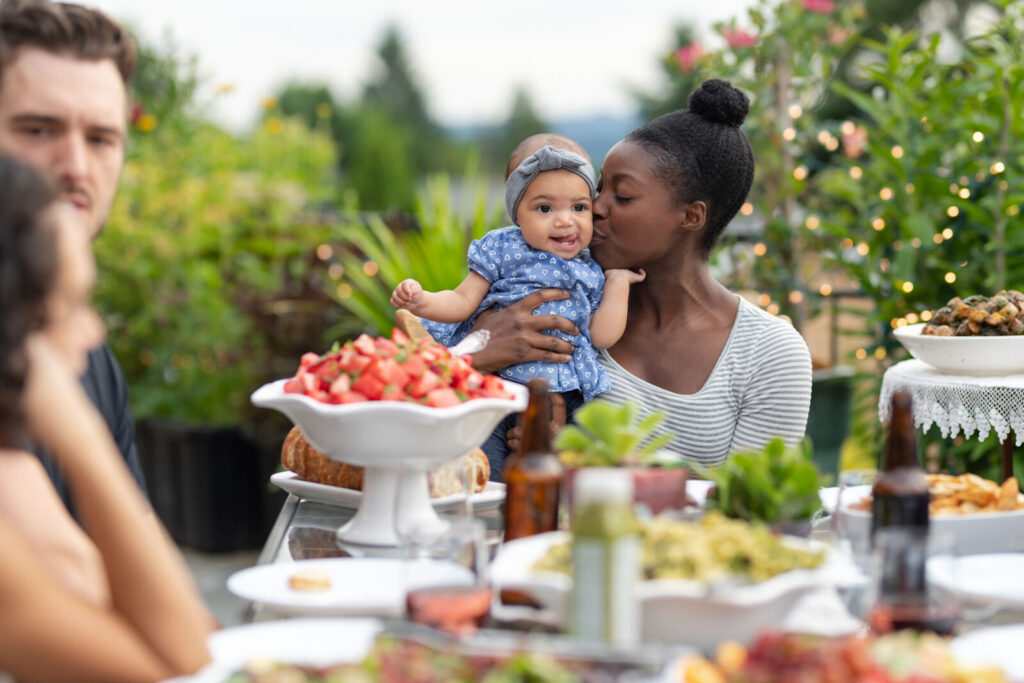 Adult holding a kissing a baby on the cheek while sitting outdoors at a table of healthy food. Two other adults are also at the table.