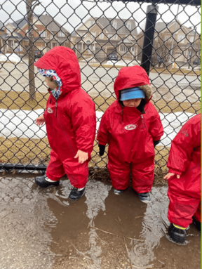 Three children wearing muddy buddy rainsuits on a rainy day in daycare yard