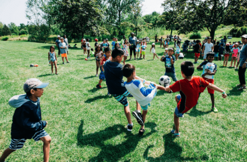 Large group of children playing a field; some are kicking a soccer ball