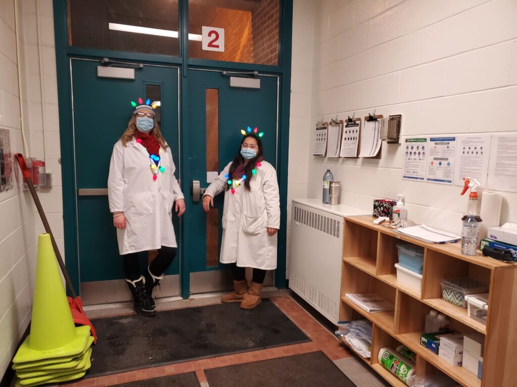 Educators standing at the centre doors during COVID-19 pandemic, wearing lab coats, medical masks, and holiday lights headbands and necklaces, 