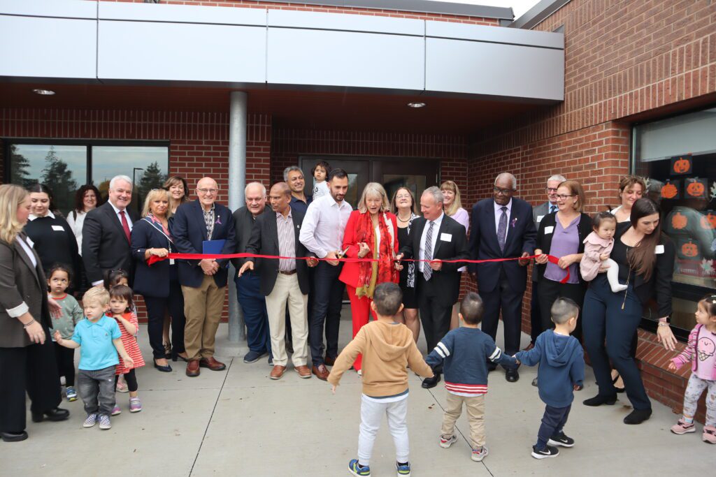 Ribbon cutting ceremony for Upper Canada Child Care at St. Rene Goupil-St. Luke CES: a group of adults, smiling, and small groups of children holding hands with each other and with staff. 