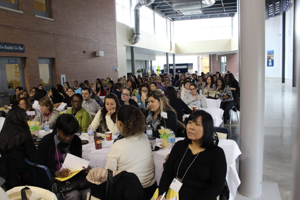 Upper Canada Child Care Professional Development Conference attendees in a large educational setting, sitting at rows of tables, observing and taking notes. 