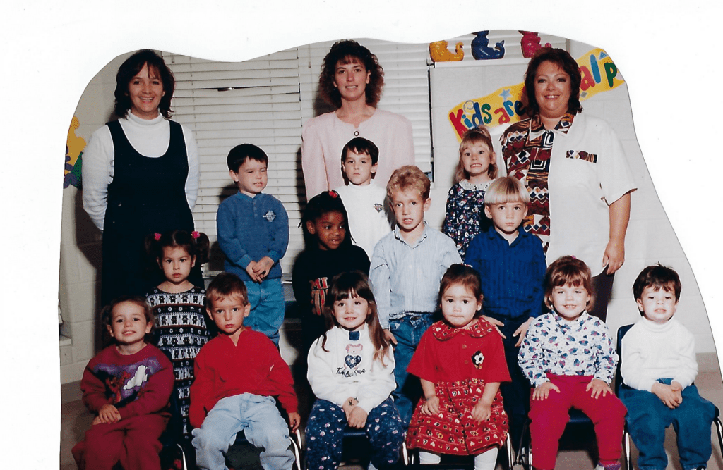 Childcare centre class photo from past decades (1993-2010). Three educators standing in the back row. Front row of children sitting and two rows of children standing.