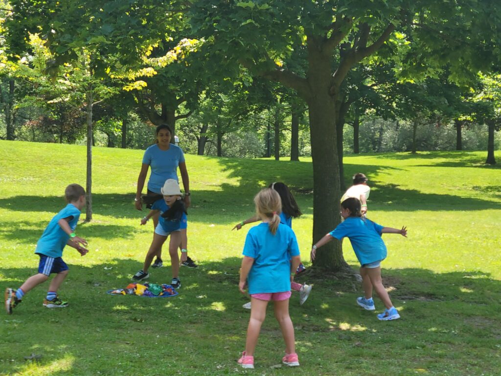 A group of 5 children playing a game with a camp staff, in a park by a tree