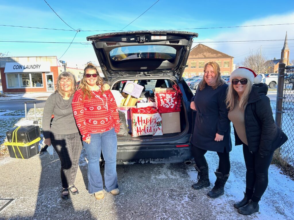 Four adults standing beside an open car trunk full of food donations on a sunny day