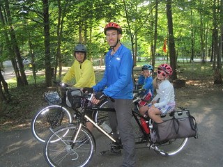 Two adults and two children smiling and standing with bicycles outside in a park with lots of trees - each bike has an adult at the front and a child on the back