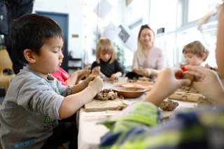 Young children at a table focused on molding clay, with adult in the background