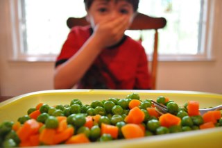 Child sitting on a chair with a plate of peas and carrots in the foreground - child covering their mouth appearing to not like the food
