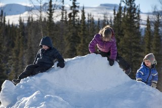 Three school age children climbing on a snow hill with evergreen trees and mountains in the background