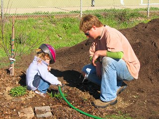 Child and adult crouched down in a garden with big pile of dirt; child holding a garden hose