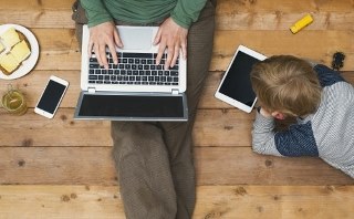 Adult on a laptop sitting on the floor beside a child that is lying on the floor with a tablet. There is food and a phone beside the adult.