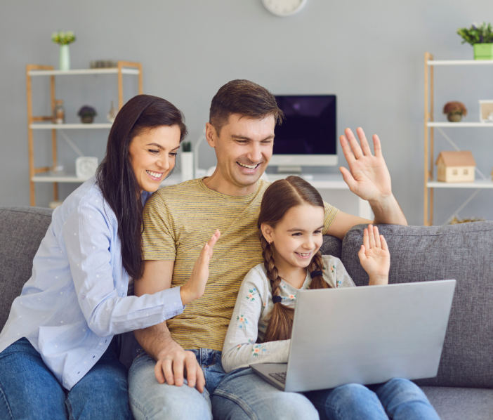 Two adults and a child sitting on a couch, smiling and waving to someone on a computer 