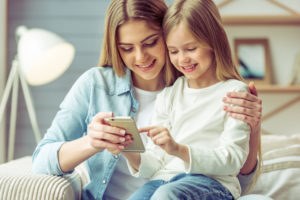 Adult and child sitting together looking at phone. Adult's arm is around child. Both are smiling.