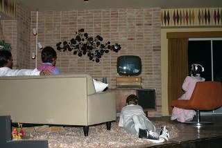 Older image of a family watching television in a living room. Parents on sofa, children on floor and chair.