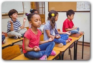 Children meditating, sitting cross-legged on top of school desks