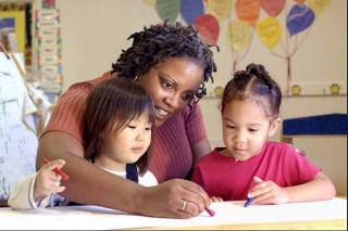 An adult with two children drawing at a table in a classroom