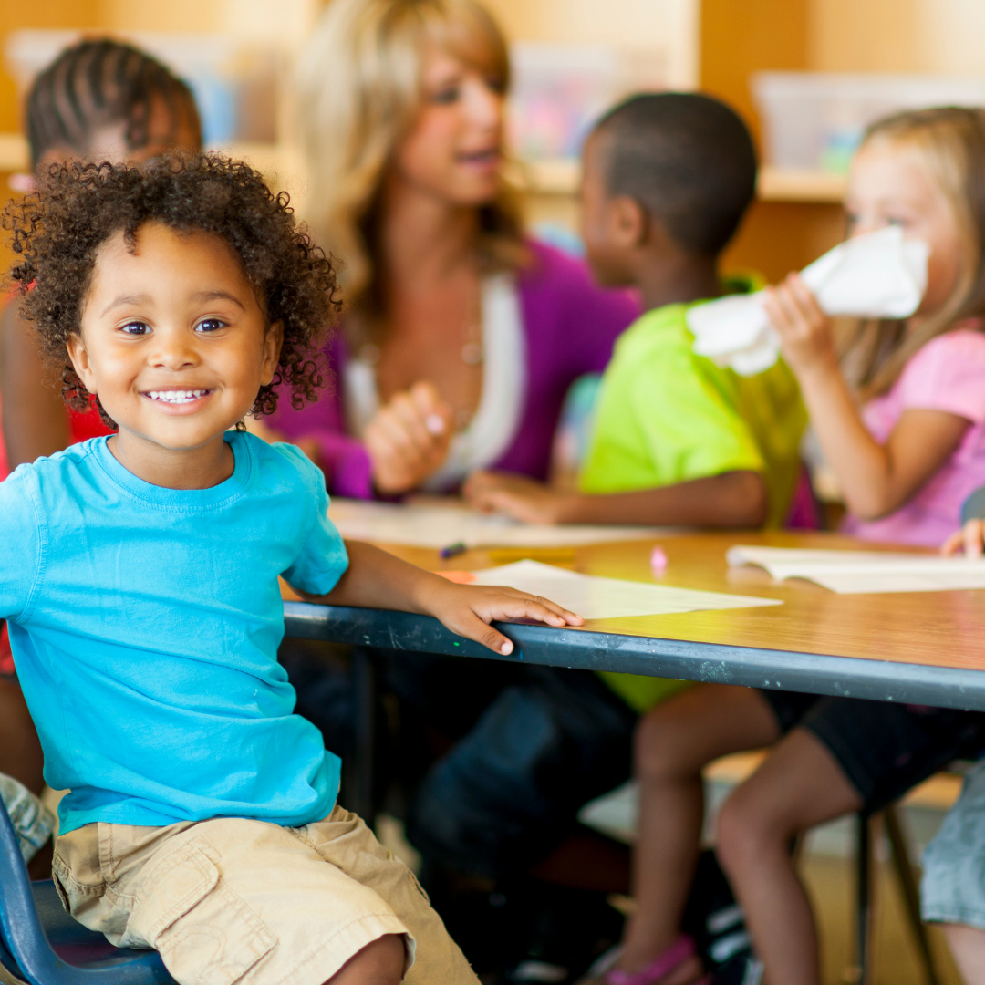 Smiling child in a preschool setting, sitting at table with other children and an adult in the background