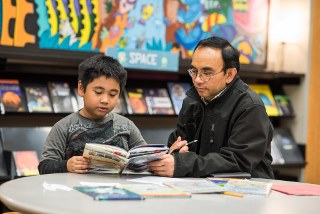 School age kid and adult sitting together at the library; child reading a book as adult looks on