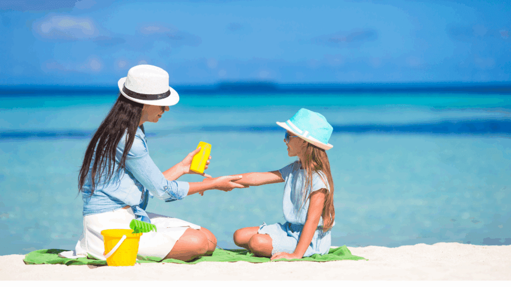 Adult and child wearing sunhats, sitting on a beach blanket, with sand bucket. Background is blue water and sky. Adult applying sunscreen to child's arm. 
