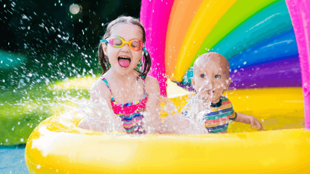 Two happy young children (older and younger toddler) sitting and splashing in a plastic wading pool. outside Half the pool has a rainbow cover.