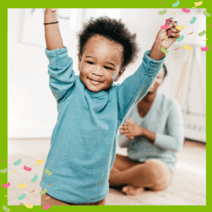 Happy toddler with arms in the air, with adult sitting in the background